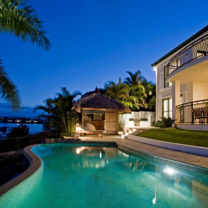 Luxurious mansion exterior at dusk overlooking pool, canal and Bali hut
